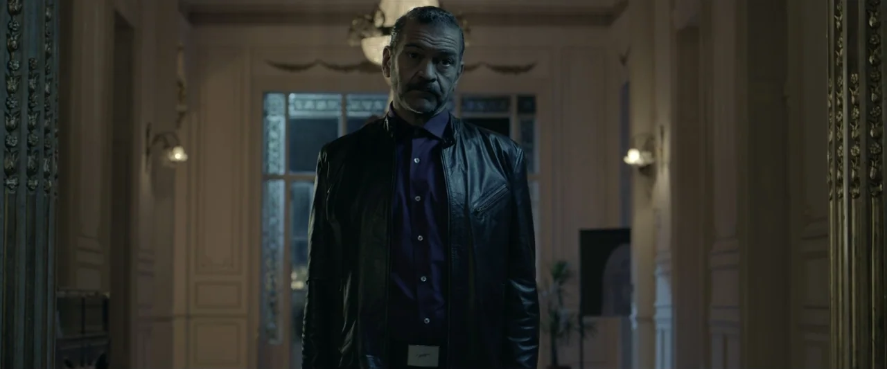 Perdida (2018) movie still: medium — A dimly lit mansion foyer with ornate wall moldings and a glass-paned double door. A man stands at the center…