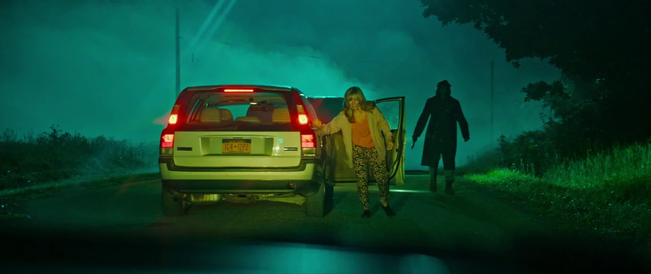 Random Acts of Violence (2019) movie still: wide — A desolate rural road at night is enveloped in a thick, artificial green fog. A woman stands…