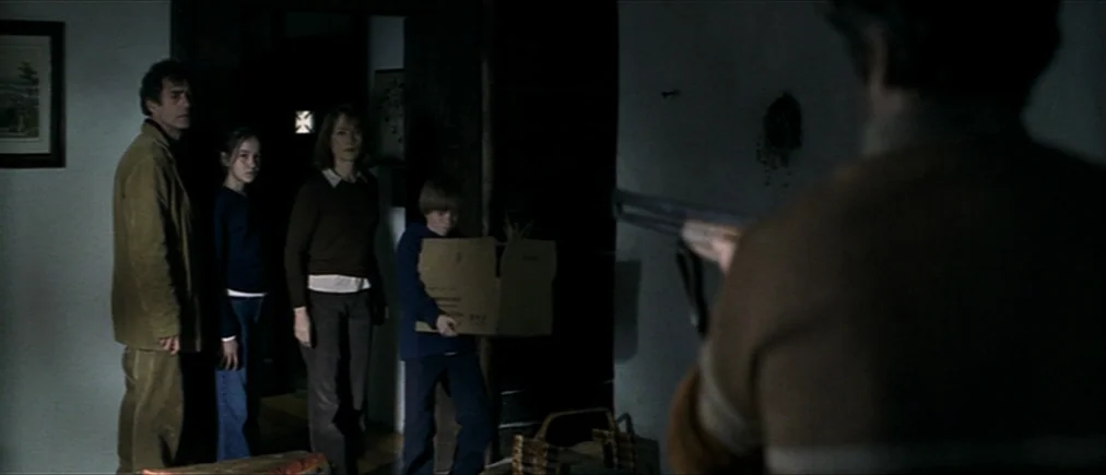 Time Of The Wolf (2003) movie still: medium — A dimly lit interior hallway with white walls and a large, dark doorway. A man aims a shotgun toward a…
