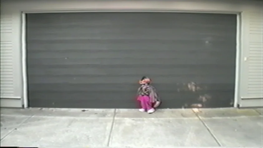 Trash Humpers (2009) movie still: wide — A concrete residential driveway fronts a large, dark-colored sectional garage door. A small child sits on…
