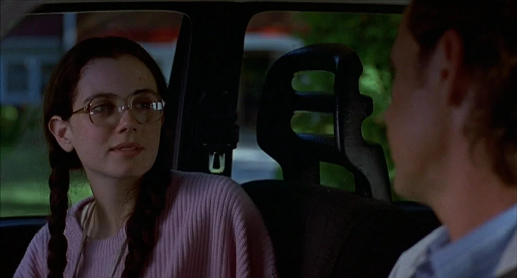 Exotica (1994) movie still: medium — The interior of a moving vehicle during the day showing the passenger and driver. A young woman with braids and…