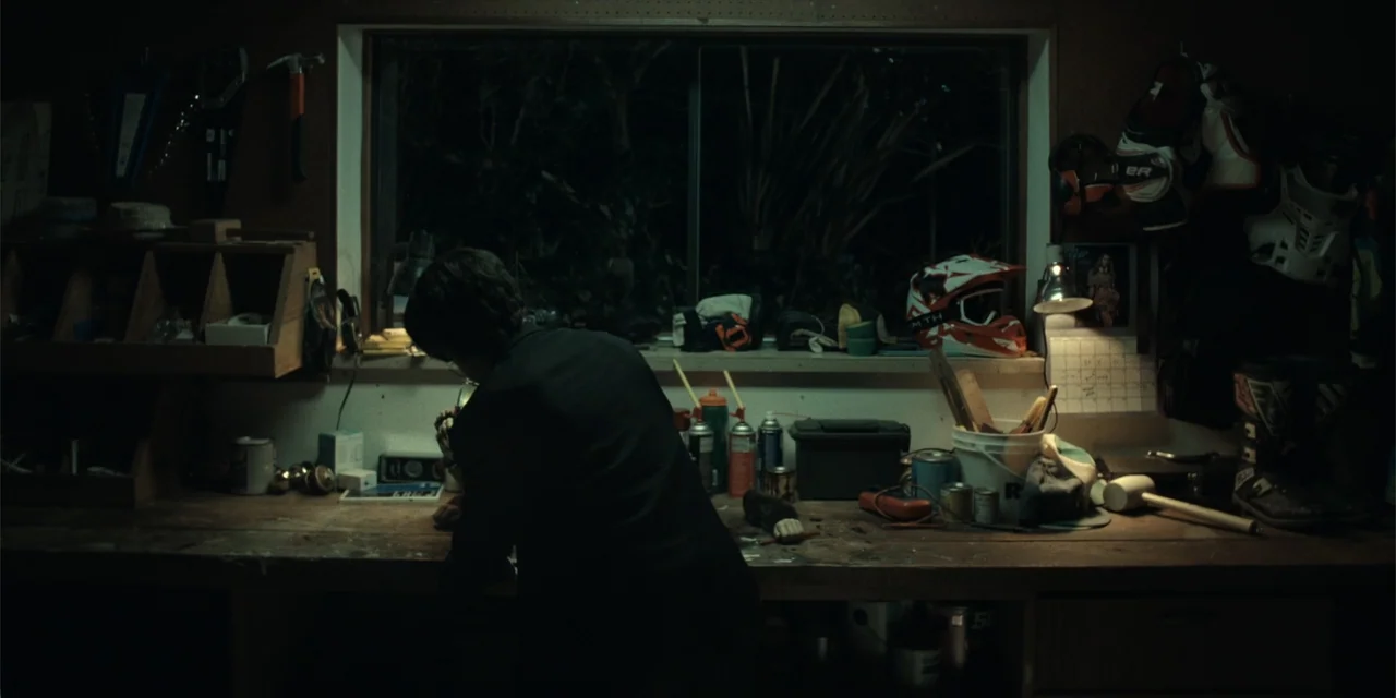 The Monkey (2025) movie still: medium — A dark workshop contains a workbench cluttered with tools, gear, and supplies. A person in a black shirt…
