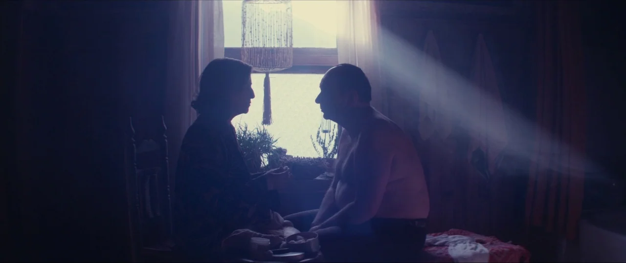 Neruda (2016) movie still: medium — A dimly lit room with a single window acting as the primary light source. Two people sit facing each other in…