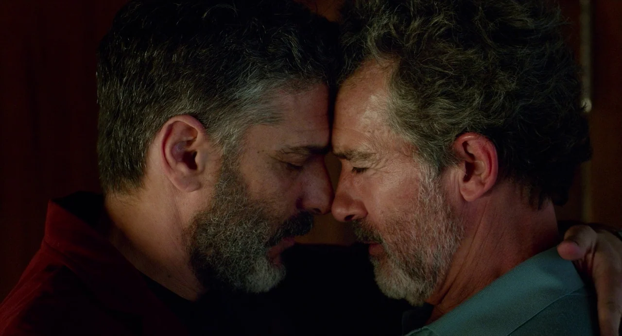 Pain and Glory (2019) movie still: closeup — A dimly lit interior space with a dark wood-paneled wall backdrop. Two middle-aged men press their…