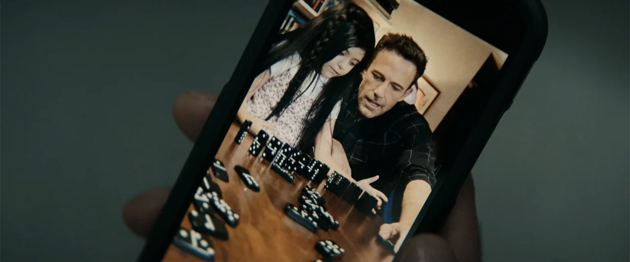Hypnotic (2023) movie still: closeup — A man and a young girl play a game of dominoes on a wooden table. The frame is a digital screen capture…