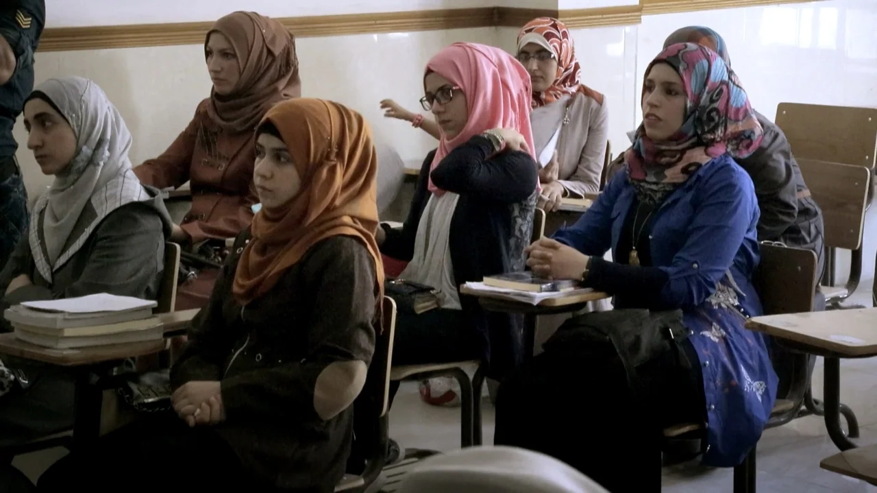 City of Two Springs (2019) movie still: medium — A quiet room features rows of small wooden desks for students. Several young women wearing…