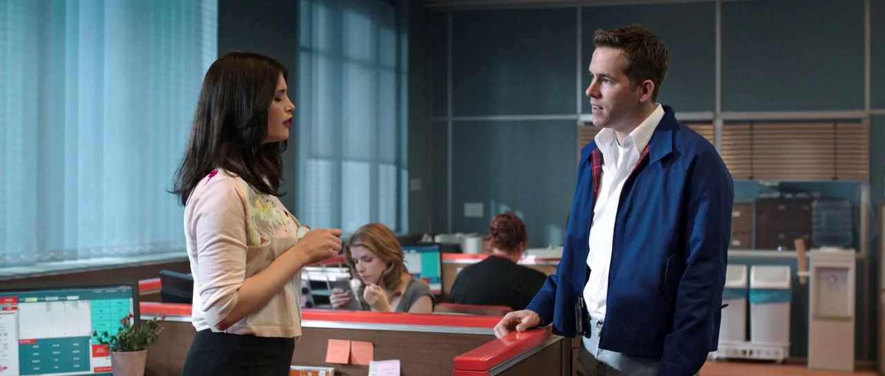 The Voices (2014) movie still: medium — An open plan office features grey cubicle partitions and blue-tinted windows. A woman in a cardigan and a…