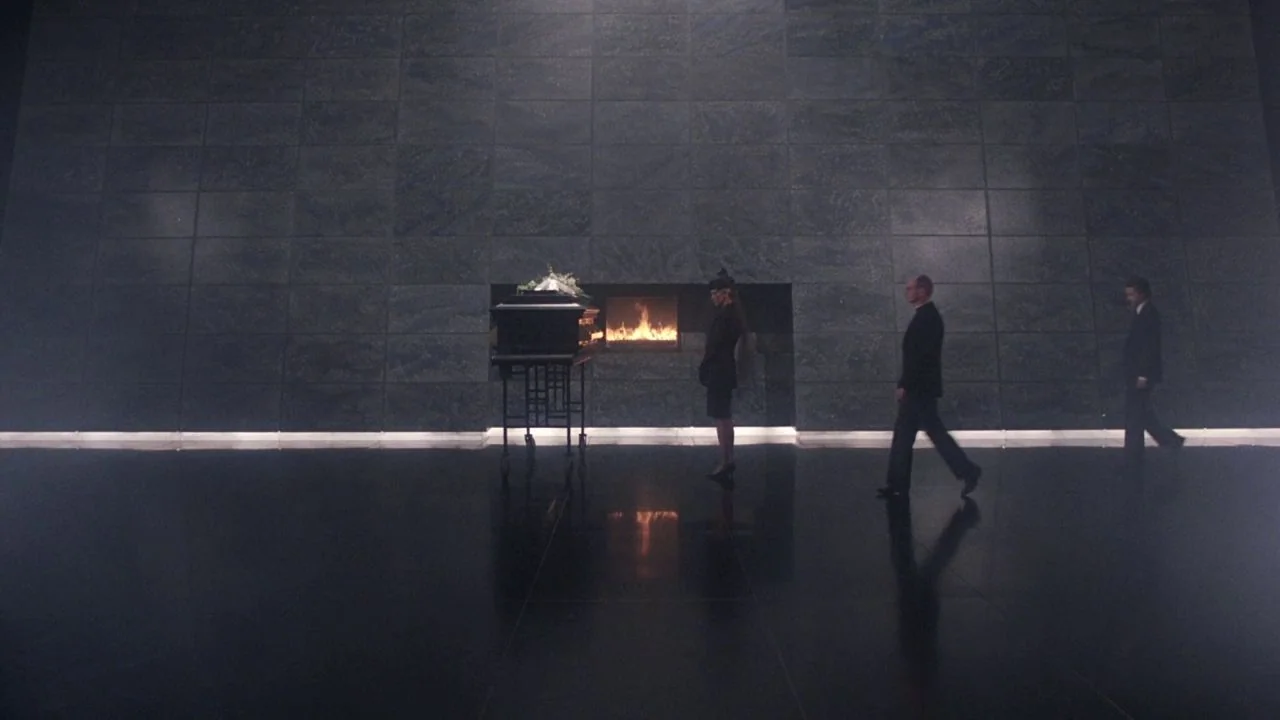 Scrooged (1988) movie still: wide — A spacious hall with large stone tiles and a central, wall-inset fireplace. Three individuals walk past a casket…