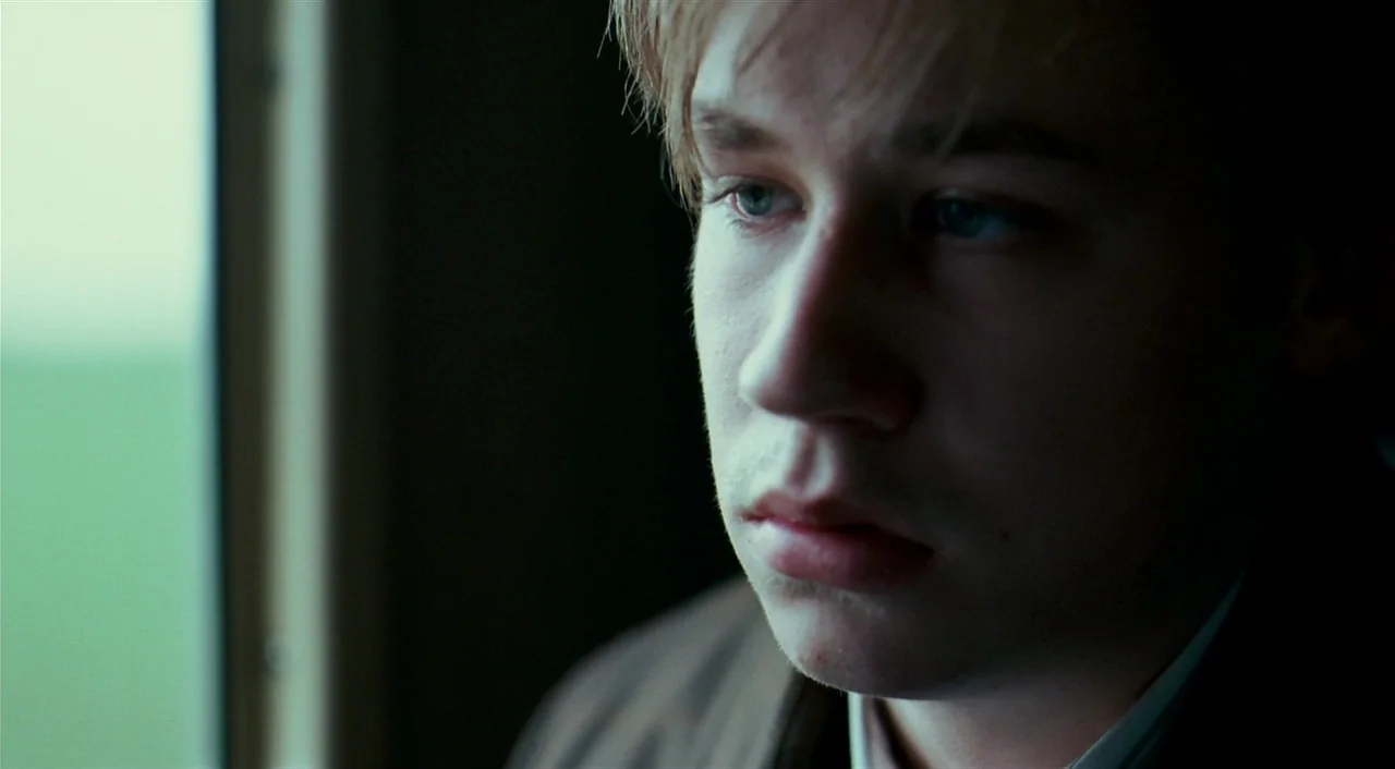 The Reader (2008) movie still: closeup — The interior of a train carriage features a window showing a blurred green exterior. A young man sits by…