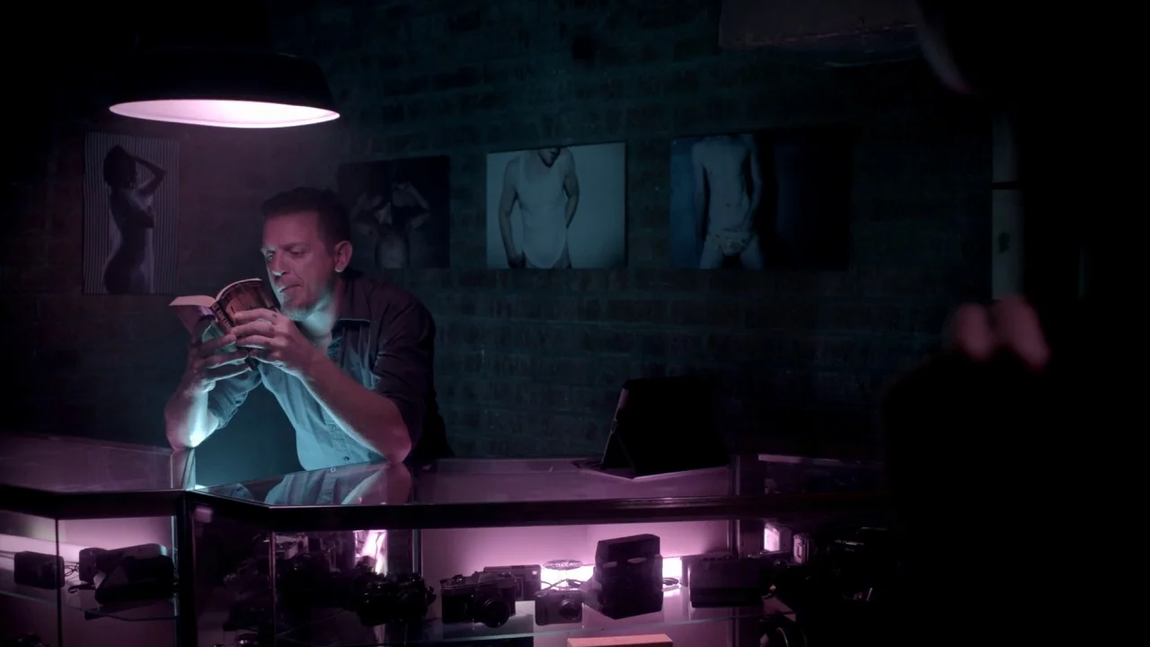 The Pact II movie still: medium — A man stands behind a glass counter in a dark, brick-walled shop filled with cameras. He leans forward under a…