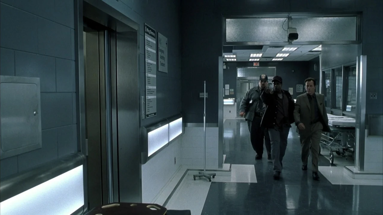 John Q (2002) movie still: fullbody — A sterile hospital corridor features grey tiled walls, a metal elevator door, and signage. Three men walk…