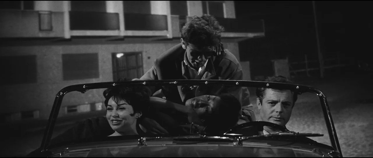 La Dolce Vita (1960) movie still: medium — A night exterior shot features three people crowded into a small open convertible. A multi-story building…