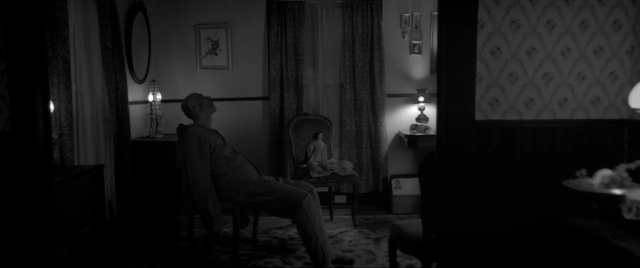 The Eyes of My Mother (2016) movie still: wide — A dimly lit vintage room features floral wallpaper and antique wood-framed furniture. A man sits in…