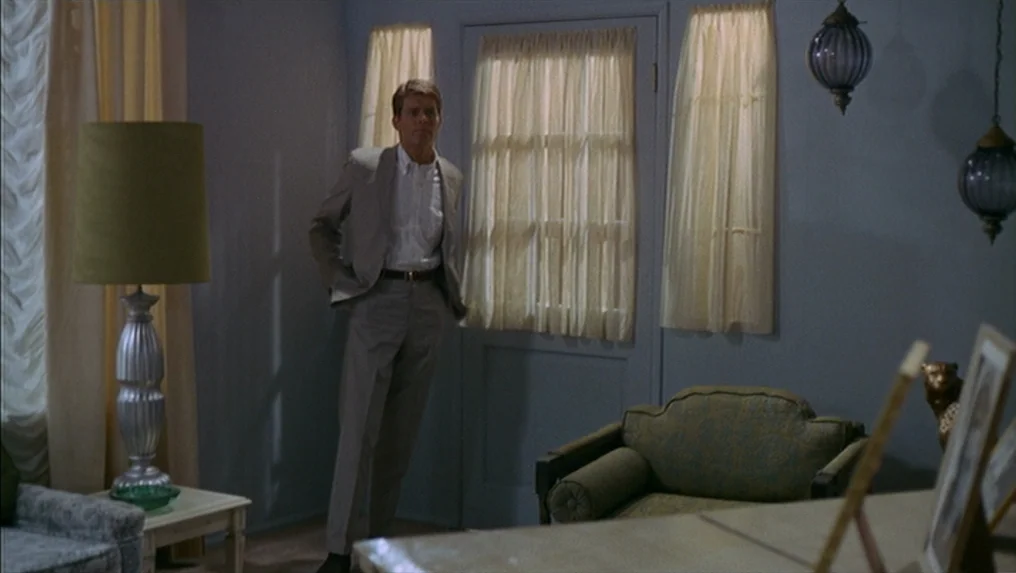 Targets (1968) movie still: fullbody — A 1970s room features pale blue walls, yellow curtains, and an ornate tall floor lamp. A man in a light grey…