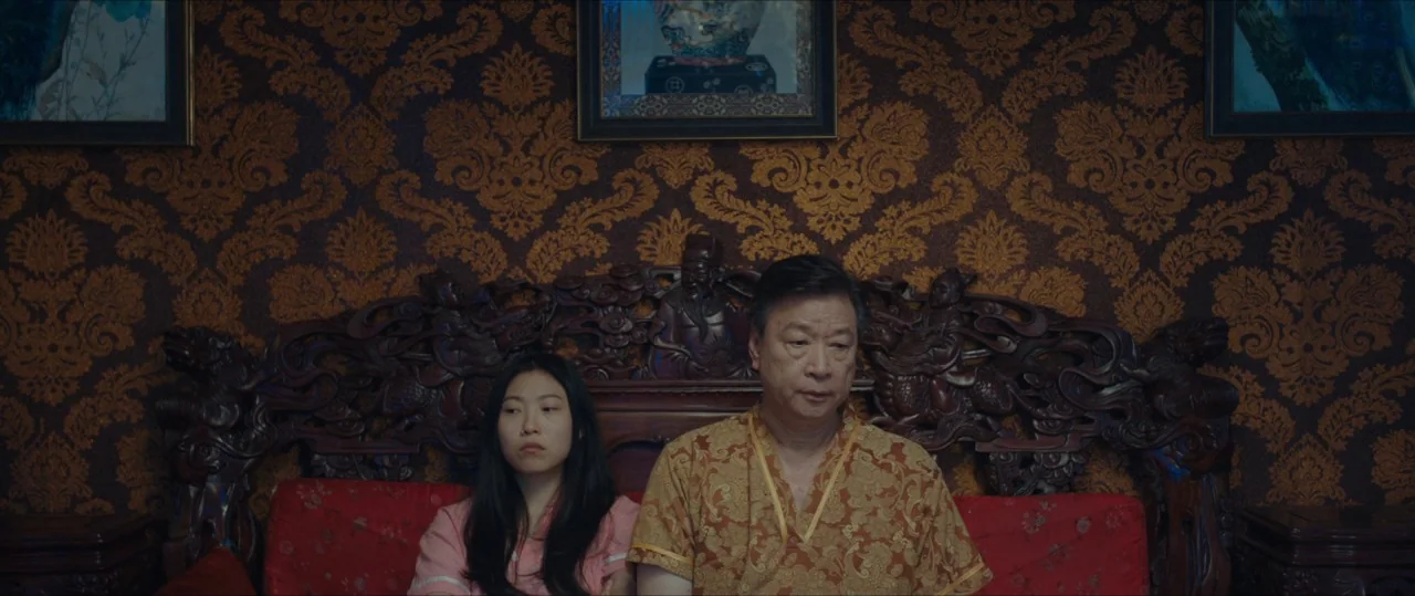 The Farewell (2019) movie still: medium — An interior space features patterned dark gold wallpaper and an elaborately carved dark wood couch. Two…