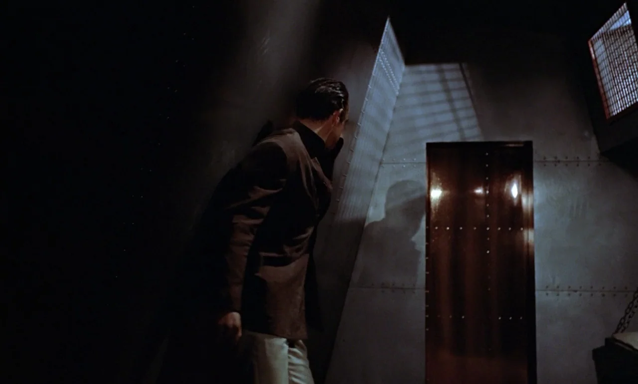 Dr. No (1962) movie still: medium — A dimly lit room features walls made of riveted metal plating. A man stands silently while observing a…