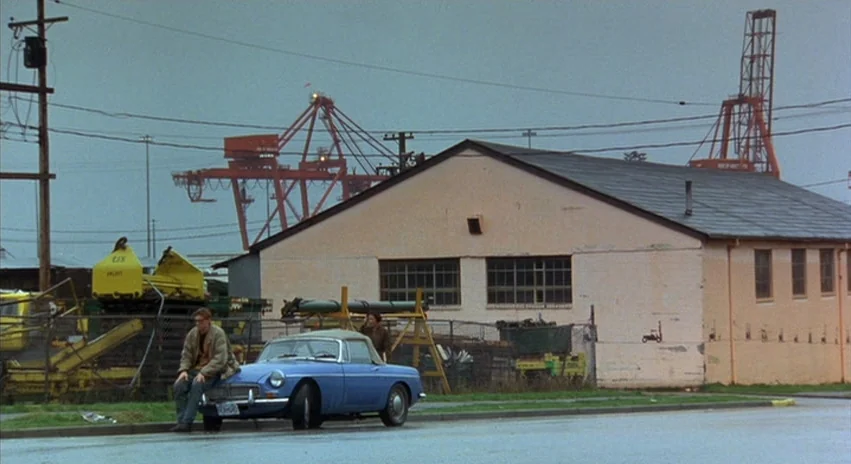 Double Happiness (1994) movie still: wide — An industrial lot features a single-story pink warehouse and large red crane structures. A blue vintage…