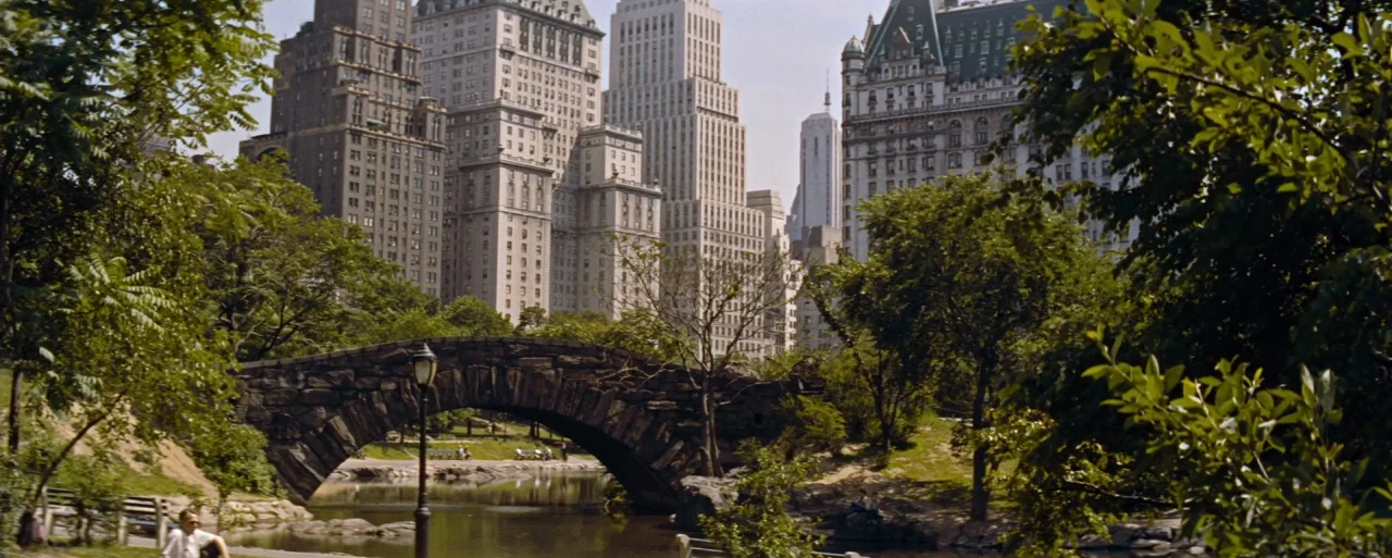 How to Marry a Millionaire movie still: establishing — A stone bridge spans a small pond surrounded by lush green trees in a park. Tall city…