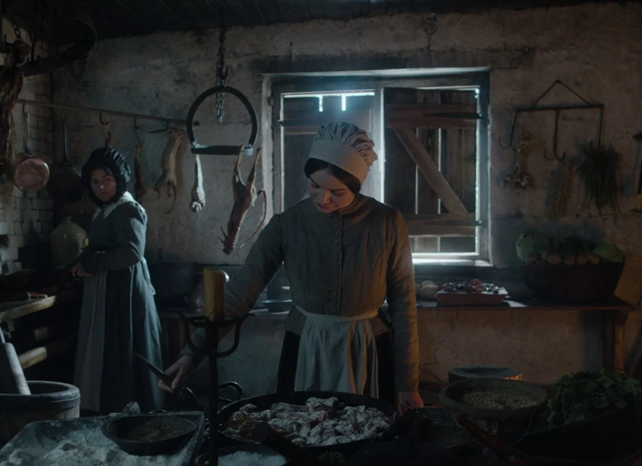 The Nightingale (2018) movie still: medium — A stone-walled pantry features rustic hanging game and assorted cooking vessels in the shadows. Two…