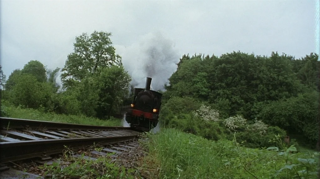 Shadow of the Vampire (2000) movie still: wide — A dark steam locomotive approaches on a curved track through a lush green landscape. Dense trees…