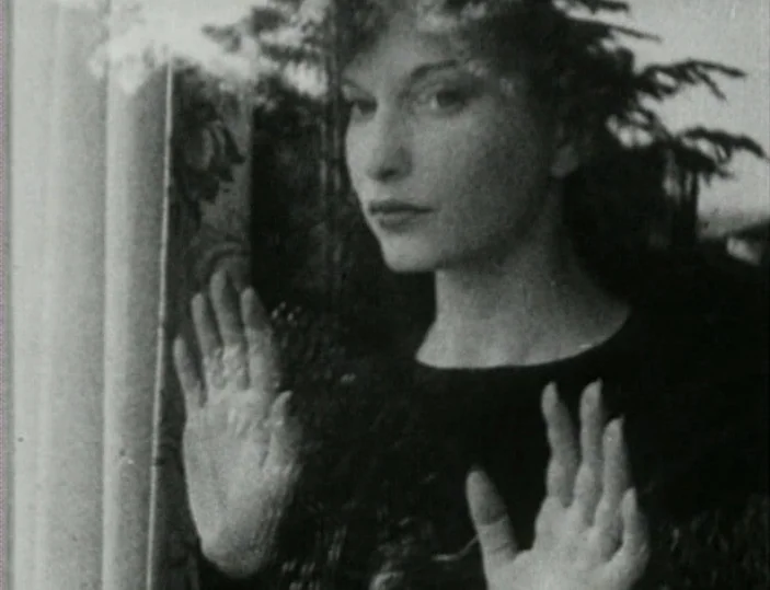 Meshes of the Afternoon (1943) movie still: closeup — A close-up shot of a woman looking through a window with trees visible outdoors. Her hands are…
