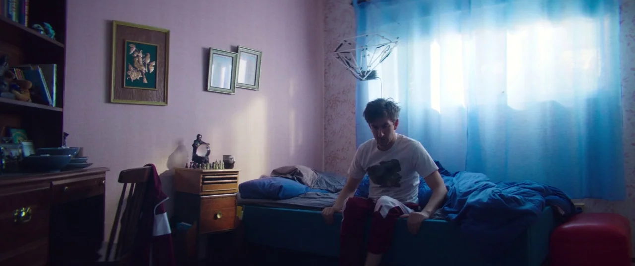 Room For Rent (2017) movie still: medium — A small room features light purple walls and a single bed with blue bedding. A man sits on the side of…