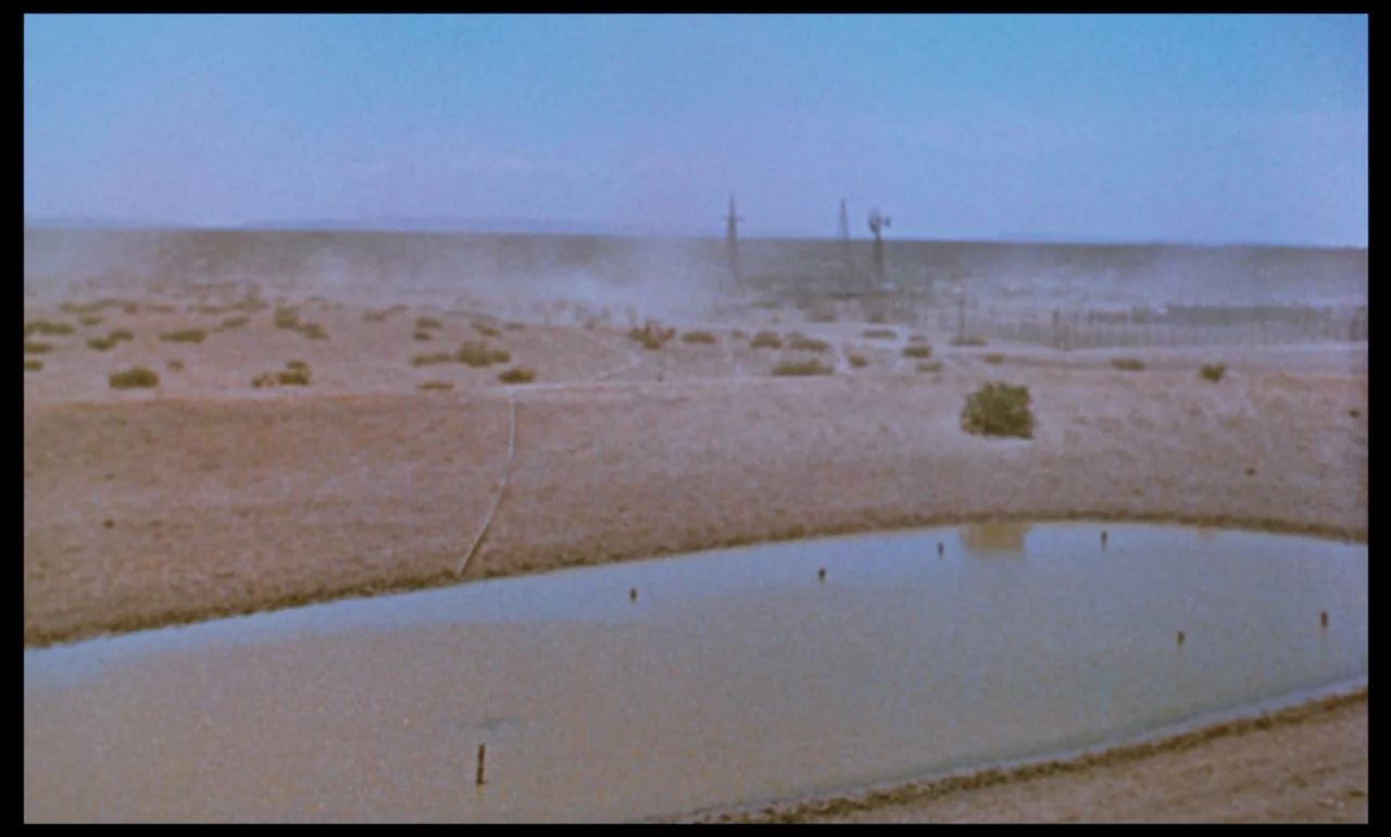 Giant (1956) movie still: establishing — A wide view of a dry desert landscape features a small muddy watering hole. A fence and distant windmills…