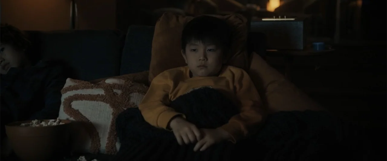 Afraid (2024) movie still: medium — A dim living room features a sofa with pillows and a bowl of popcorn. Two children sit on the couch while…