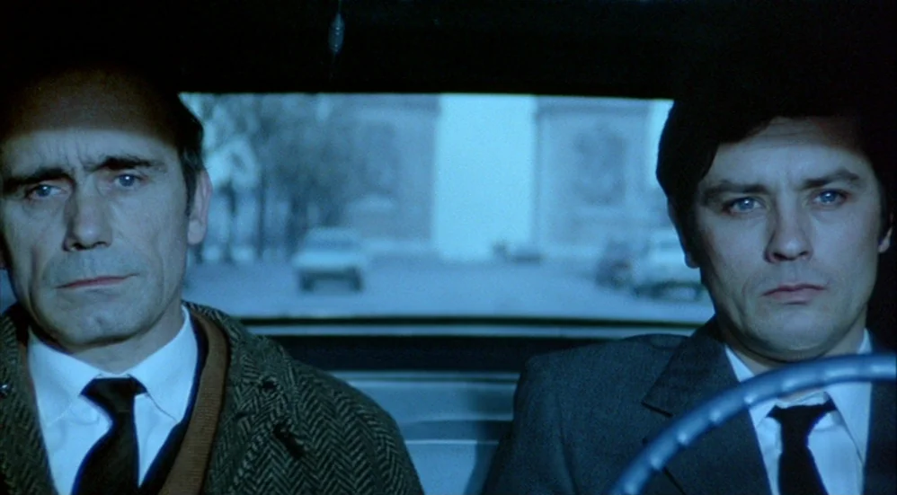 Un Flic (1972) movie still: medium — Two men in business attire sit in the front seats of a car. Through the rear window, the Arc de Triomphe is…