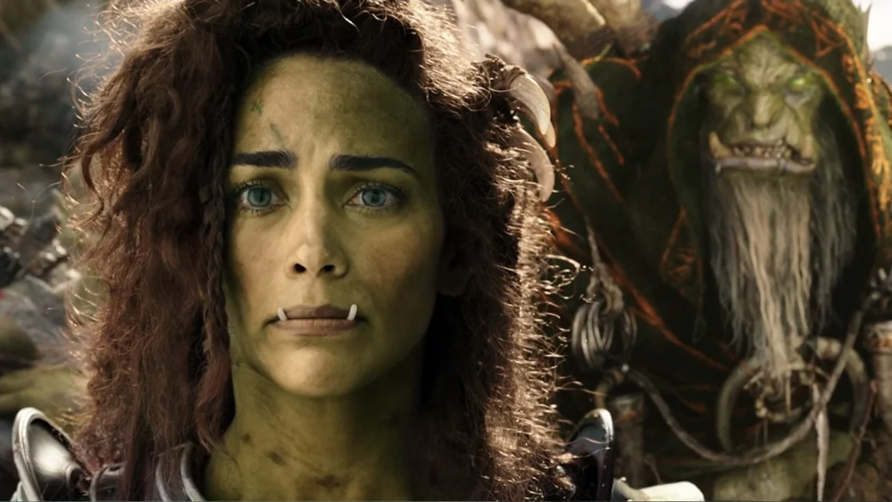 Warcraft (2016) movie still: closeup — A green-skinned woman stands in the foreground on a dusty outdoor battlefield. An elderly orc with a long…