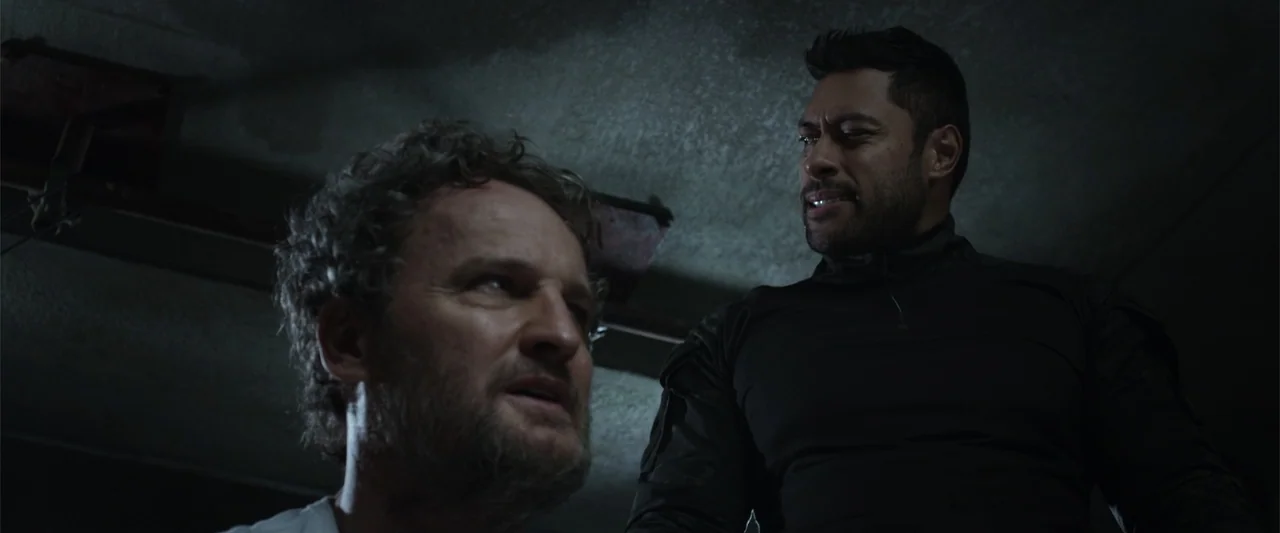 Black Site (2022) movie still: medium — A dark concrete interior with rough walls and dim overhead lighting. A man stands over a second seated man…