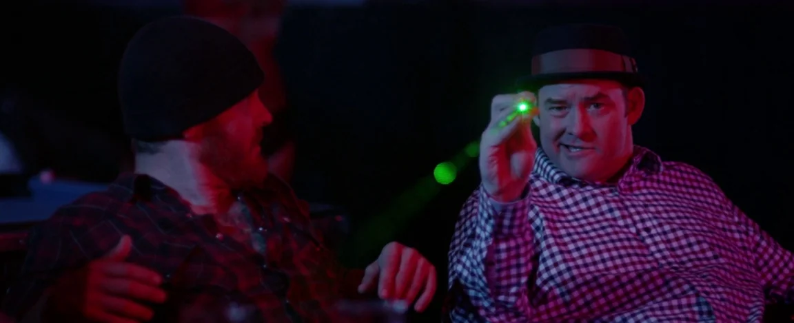 Cheap Thrills (2013) movie still: medium — A dimly lit room with red and blue light filling the space. One man holds a bright green laser pointer…