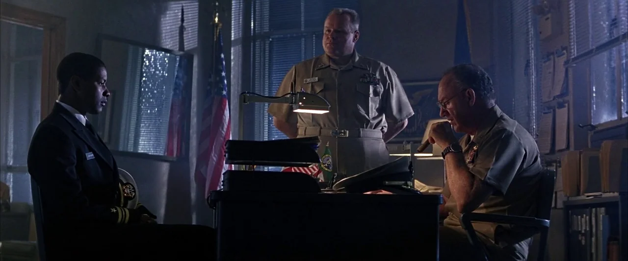Crimson Tide (1995) movie still: wide — A dimly lit office features vertical blinds, an American flag, and a cluttered desk. Three men in military…