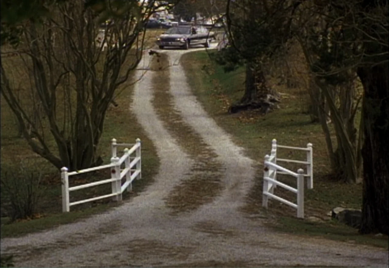 Ambushed (1998) movie still: establishing — A gravel driveway bordered by trees leads from a white gate toward a car. A dark sedan travels along the…
