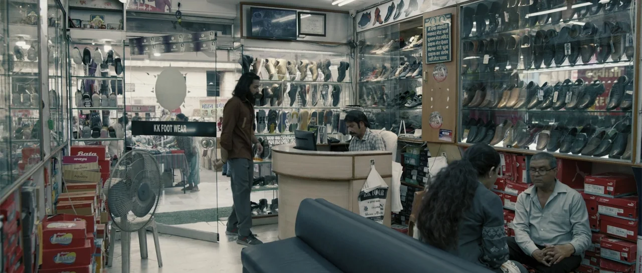 Nishaanchi 2 (2025) movie still: medium — A cluttered shoe store features glass display cases filled with various footwear. A man walks through the…