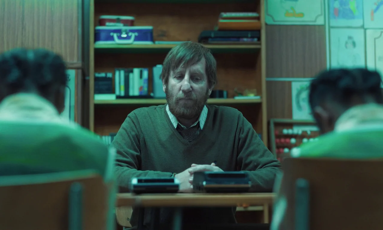 The Silent Twins (2022) movie still: medium — A classroom features wooden shelving units filled with books and various colorful school supplies. A…