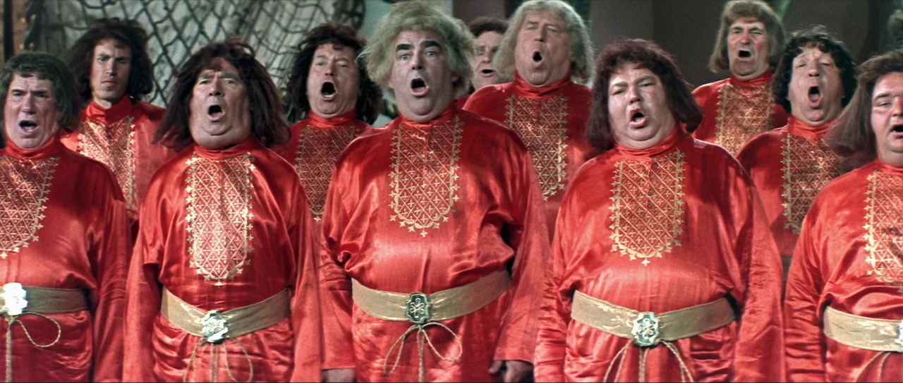 Through Fire, Water and… Brass Pipes (1968) movie still: medium — A group of men in red tunics stand side-by-side on a stage. They are singing with…
