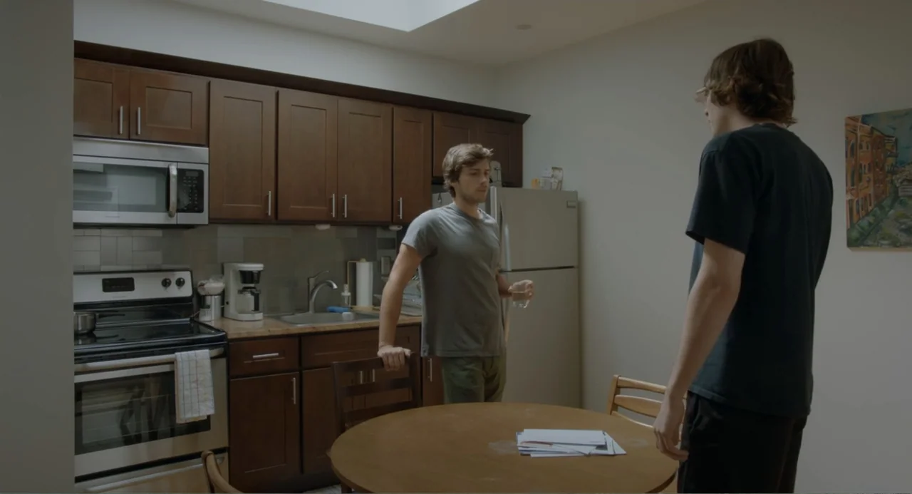 This Closeness (2024) movie still: medium — A modest apartment kitchen contains wooden cabinetry, stainless steel appliances, and a round table. One…