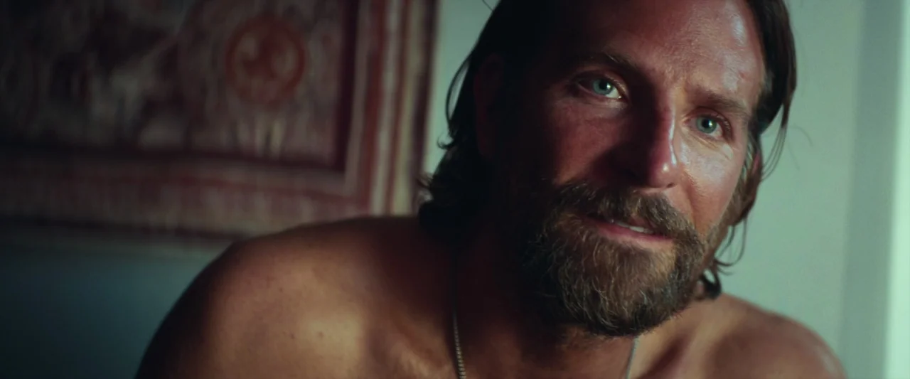 A Star Is Born (2018) movie still: closeup — A man stands in an indoor space with a textured patterned wall hanging. The man looks slightly…