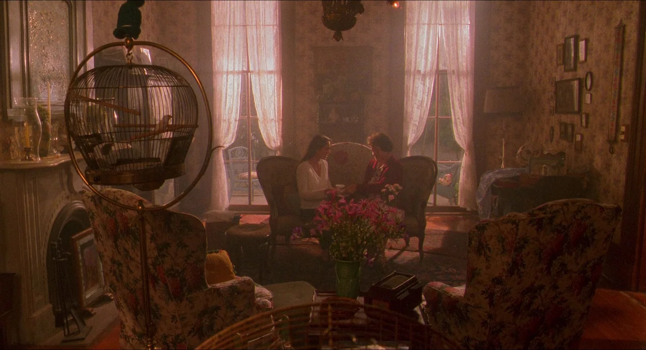 Hard Target (1993) movie still: wide — A cluttered 1990s living room features floral-patterned chairs and wallpaper with lace-curtained windows. A…