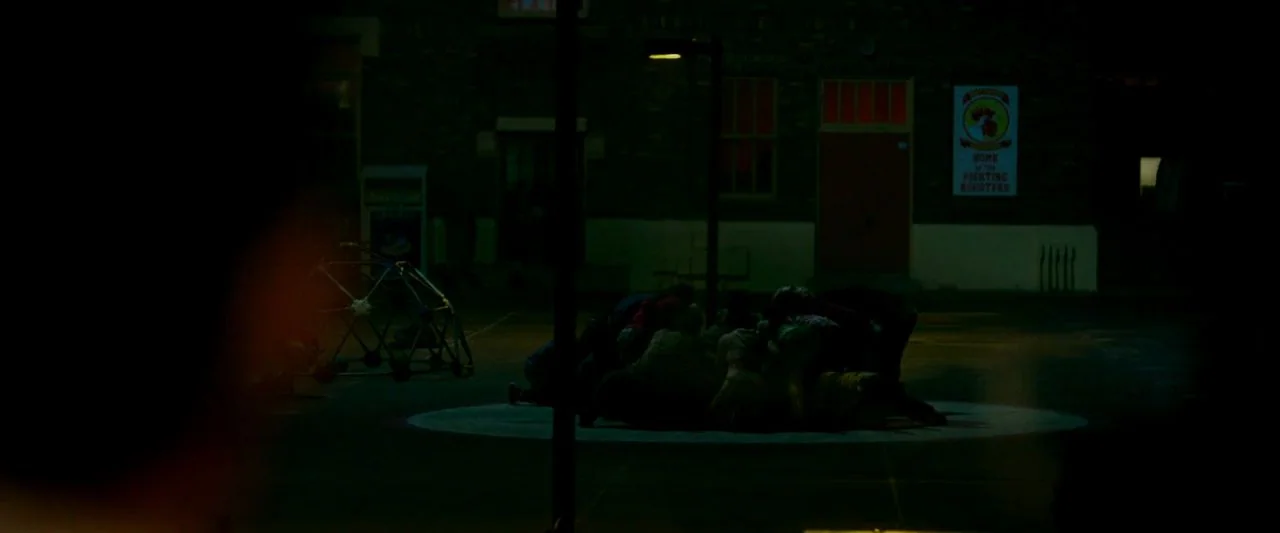 Cooties (2014) movie still: wide — A dark playground at night features a brick building with a red doorway. A small group of people huddle together…