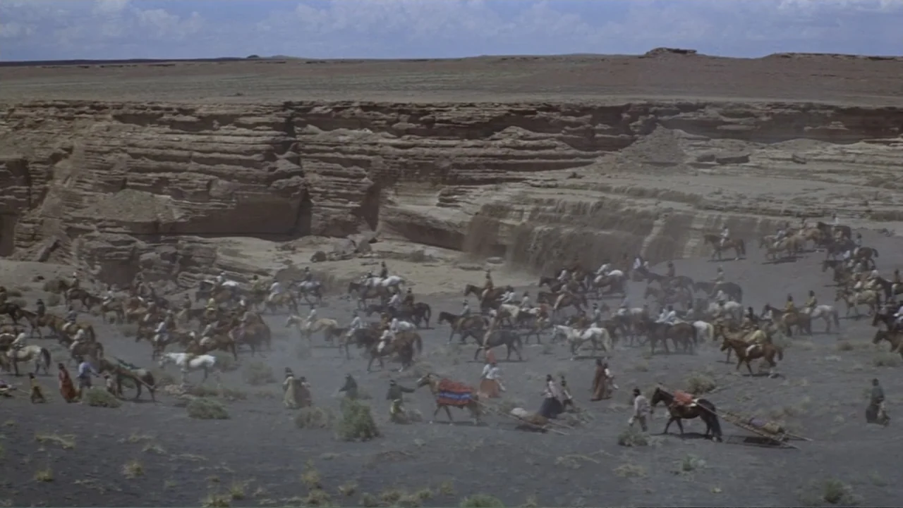 A Distant Trumpet (1964) movie still: wide — A wide shot shows a vast expanse of desert floor beneath sheer rock cliffs. A large group of people on…