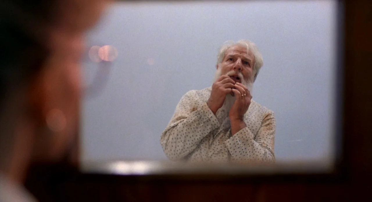 From Beyond (1986) movie still: medium — A man's reflection is centered within a framed mirror in a bathroom setting. He stands still while looking…