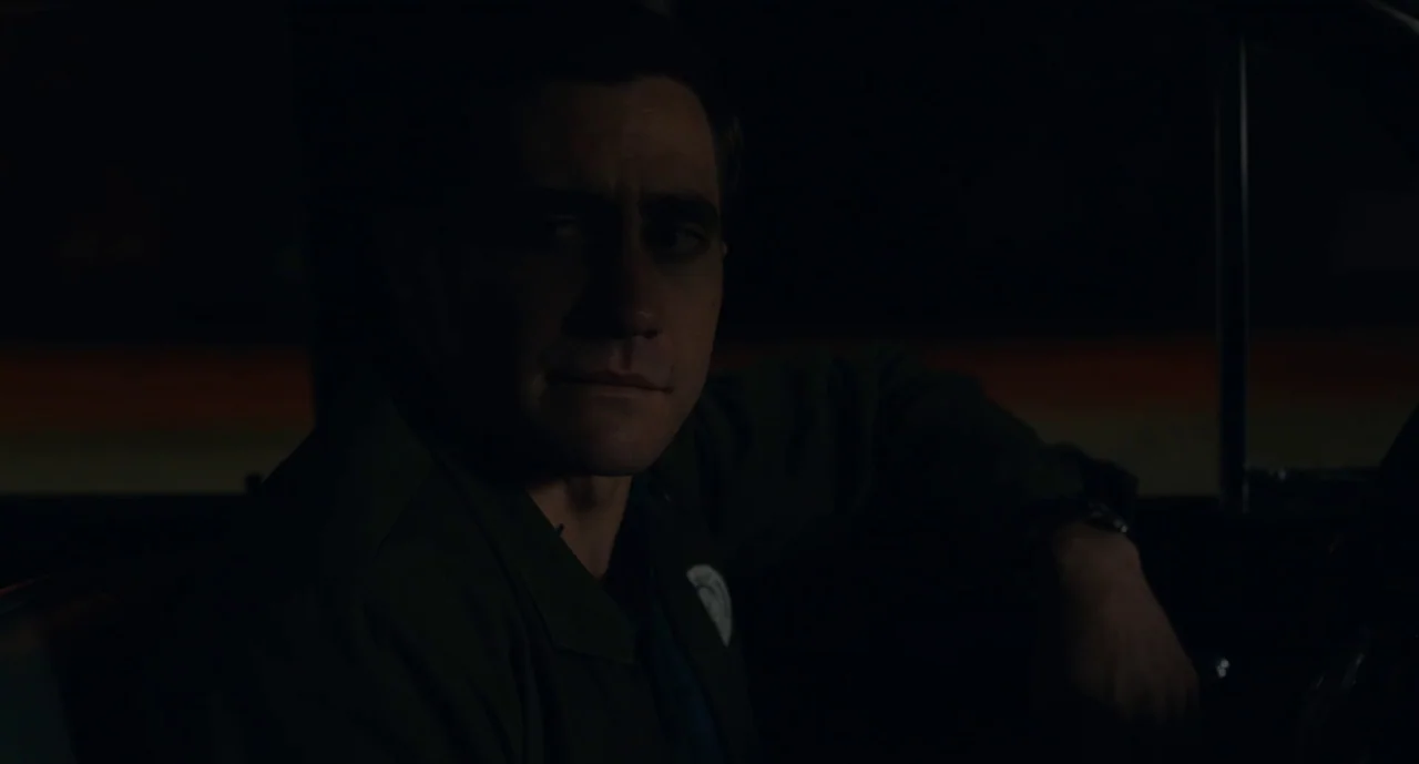 Wildlife (2018) movie still: medium — The interior of a car at night is almost entirely consumed by deep shadows. A man sits behind the steering…