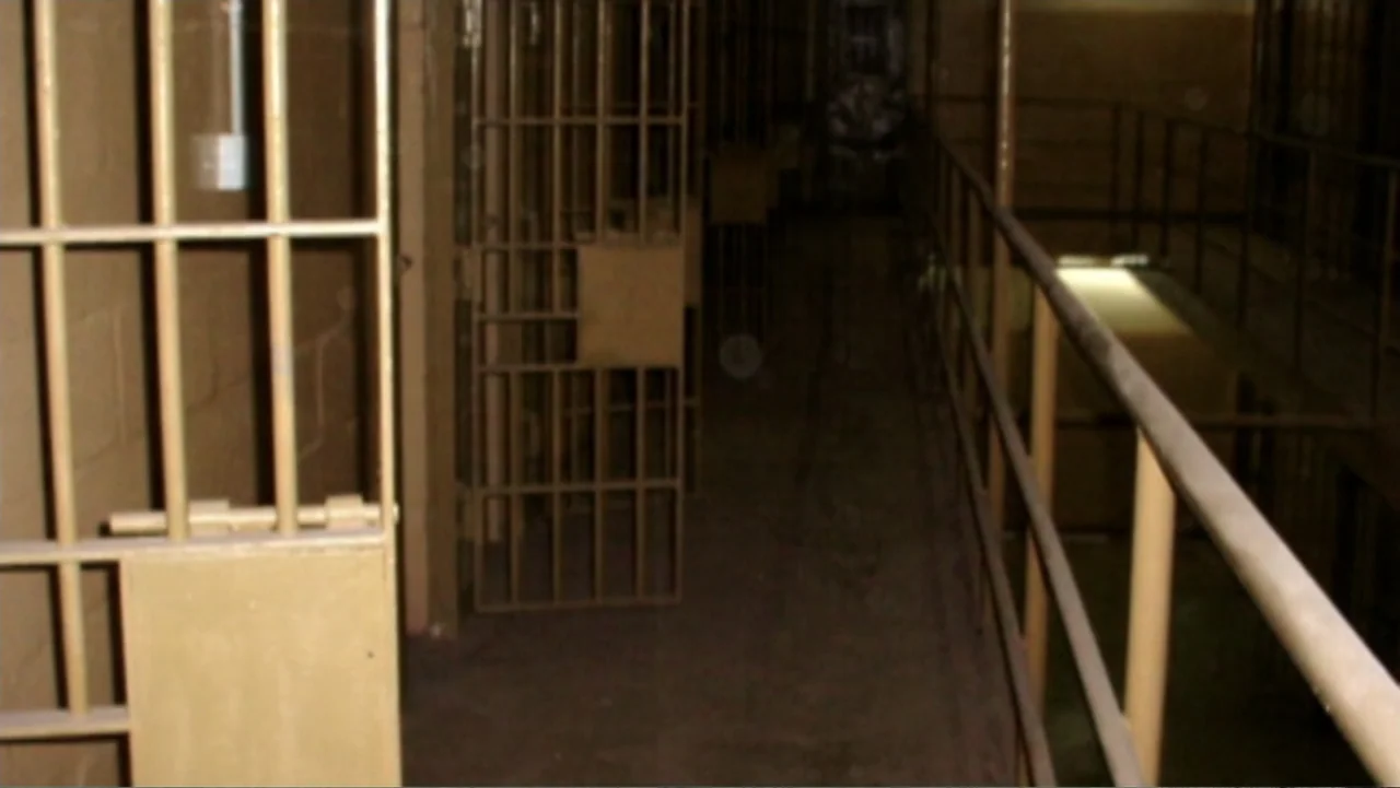 Ghosts of Abu Ghraib (2007) movie still: wide — A dimly lit prison cell block corridor features multiple rusted metal barred doors. An empty hallway…