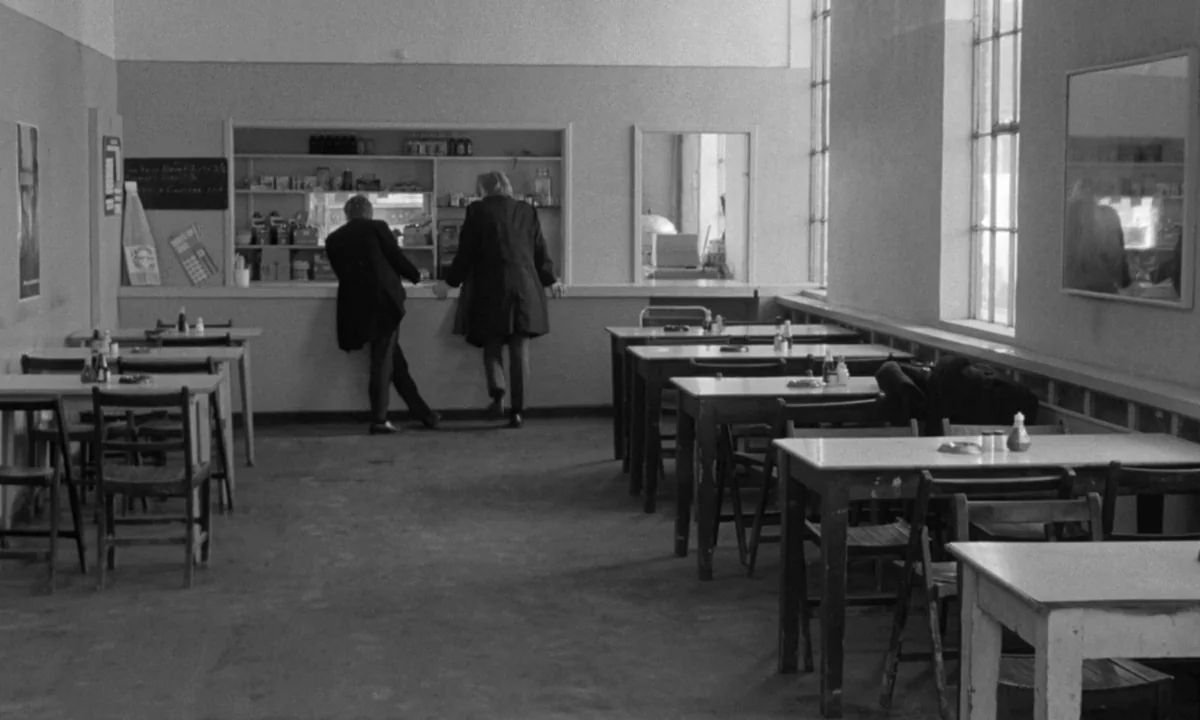 If…. (1968) movie still: wide — A sparse, institutional cafeteria with multiple wooden tables and a rear serving counter. Two men stand at the…