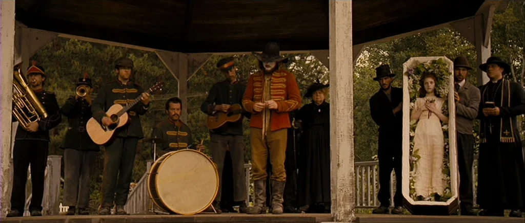 I’m Not There (2007) movie still: wide — A wooden gazebo houses a musical band alongside an upright open casket. A young girl rests inside the…
