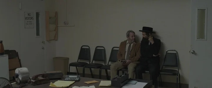 To Dust - Nightmares (2018) movie still: wide — A spare, beige-walled waiting area contains a row of black plastic chairs. Two men sit in the…