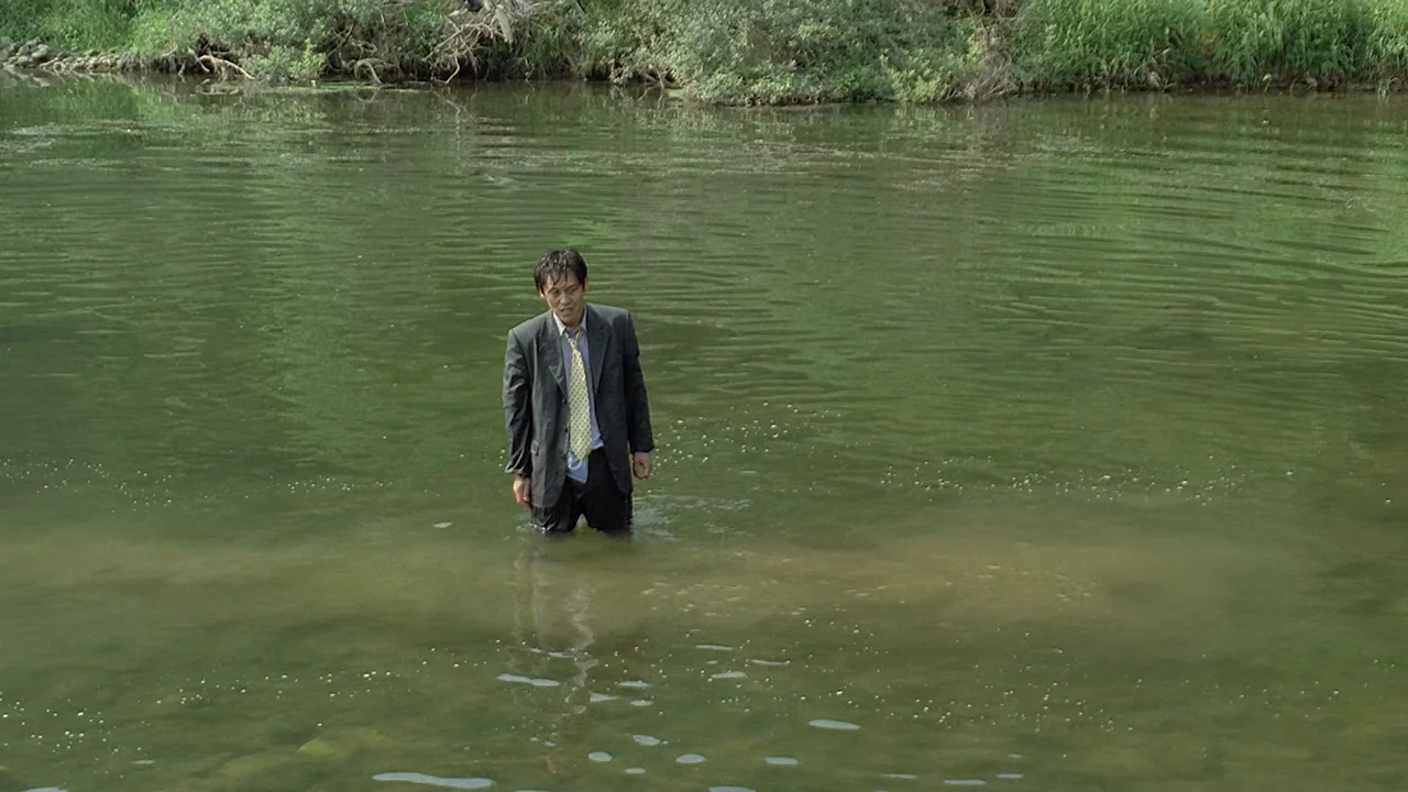 Peppermint Candy (1999) movie still: wide — A man stands in the middle of a shallow river with grassy banks. He is wearing a formal suit and is…