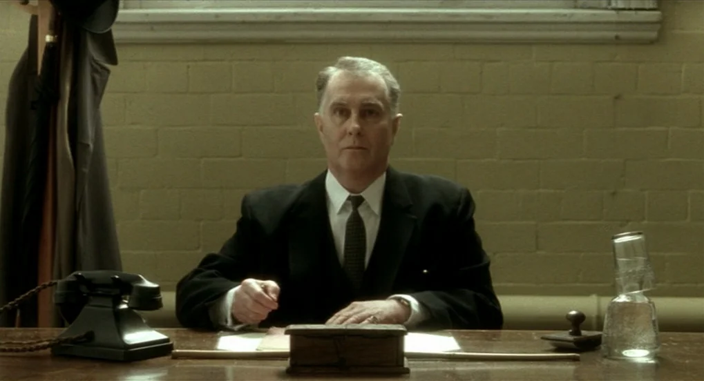 The Long Day Closes (1992) movie still: medium — A plain interior with a light-colored brick wall and a desk. An older man sits at the desk looking…